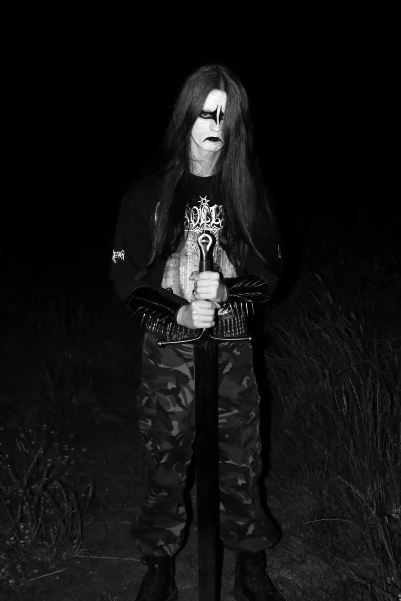 Black Metal aesthetic — This image perfectly captures the core elements: corpse paint, illegible band logo tee, spiked gauntlets, bullet belt, combat boots, and a dark, outdoor setting.