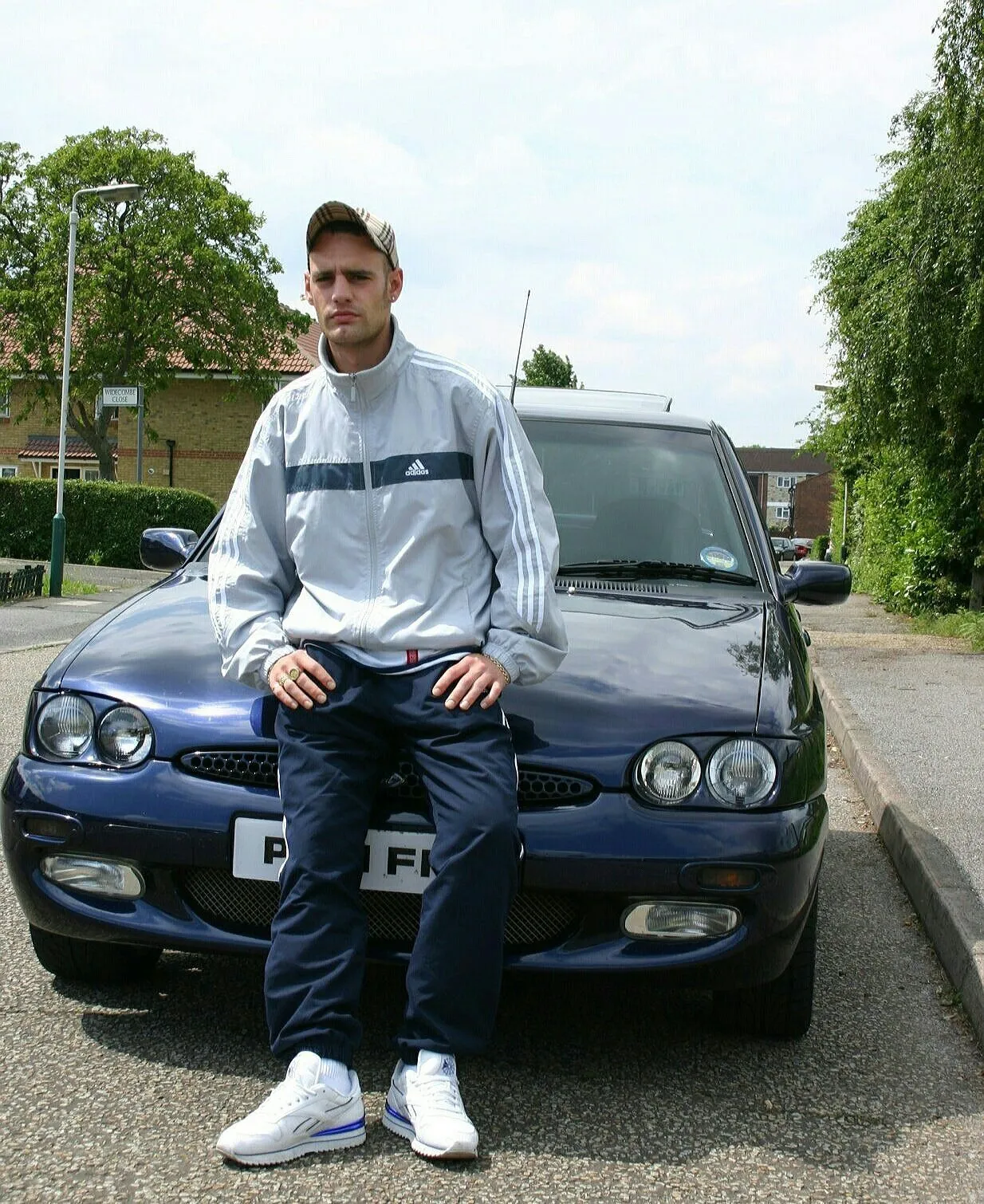 Chav aesthetic — Adidas tracksuit with tucked-in bottoms, baseball cap, and white trainers perfectly capture the described look and era.