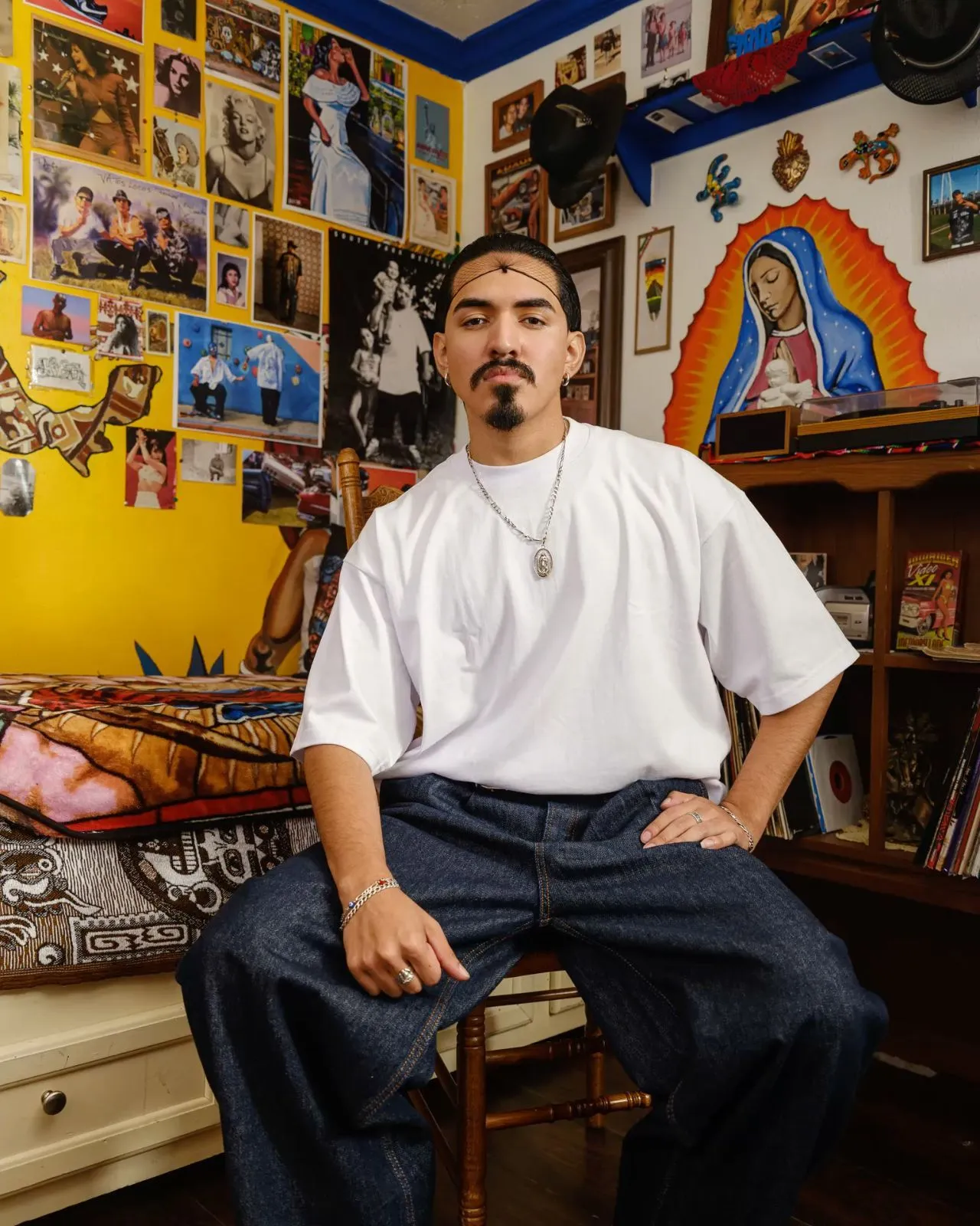 Cholo (Chicano subculture) aesthetic — The oversized white tee, baggy jeans, and the prominent Virgen de Guadalupe imagery are direct references to the described aesthetic.