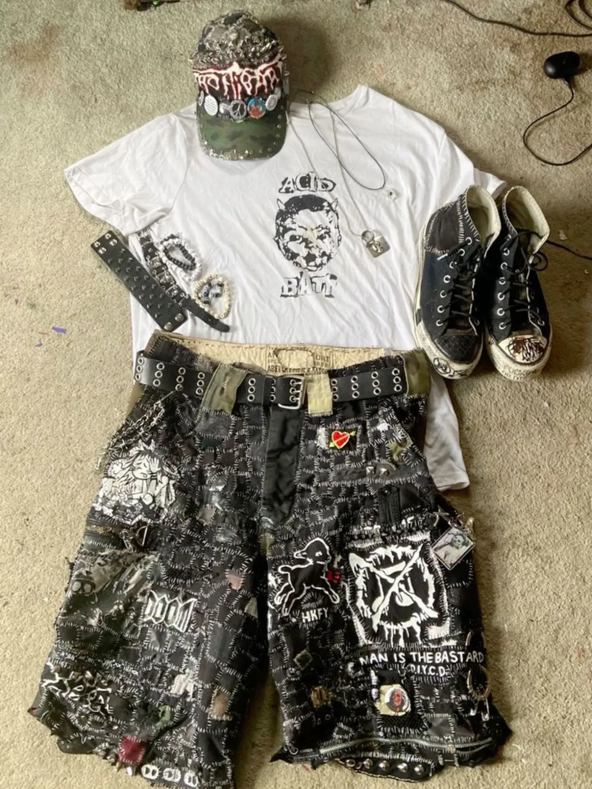 Crust Punk aesthetic — The heavily patched and studded shorts, band t-shirt, spiked wristband, and patched baseball cap are all direct references to the description's visual signature and signature garments.