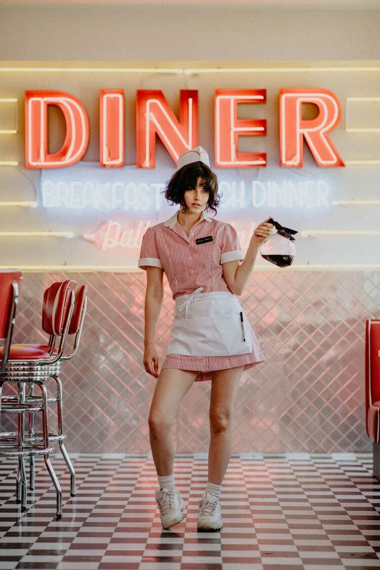Diner aesthetic — This image perfectly captures the diner waitress aesthetic with the striped shirtwaist dress, white apron, paper cap, name tag, and roller skates, all set in a diner environment.
