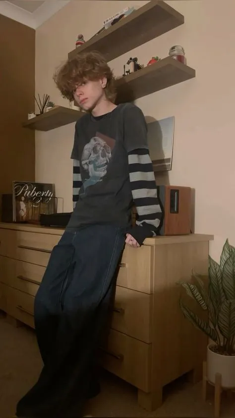 E-Boy aesthetic — The centre-parted curtained fringe, dyed hair, striped long-sleeve under a graphic tee, and baggy jeans are all direct matches to the E-Boy description's visual signature.