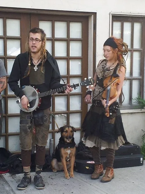 Folk Punk aesthetic — This image perfectly captures the Folk Punk aesthetic with musicians playing acoustic instruments (banjo, violin), wearing layered, thrifted-looking clothing, and a general busker/DIY vibe.