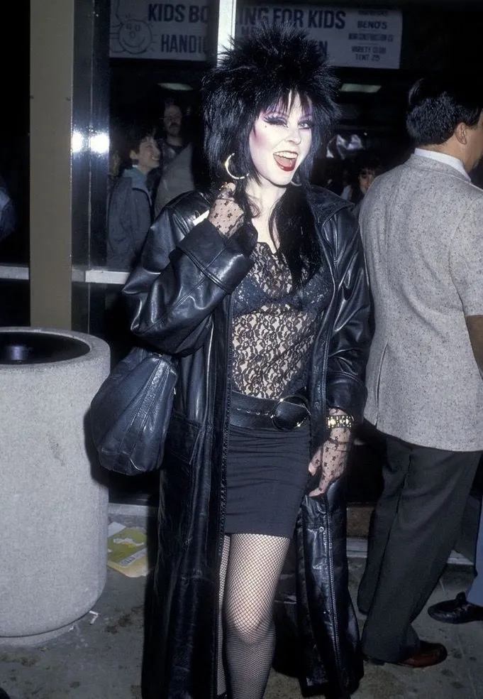 Gothabilly aesthetic — The leather trench coat, lace top, fishnets, and heavy makeup strongly evoke the deathrock/horror-punk adjacent elements and the overall dark aesthetic.
