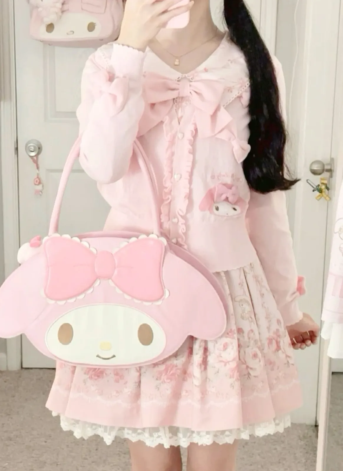 Kawaii aesthetic — The image perfectly embodies the Kawaii aesthetic with its soft pastel pink palette, rounded shapes, ruffles, bows, and character merchandise (My Melody bag and patch). The outfit includes a ruffled blouse, a floral skirt with lace trim, and a bow, all aligning with the description's visual signature and signature garments.