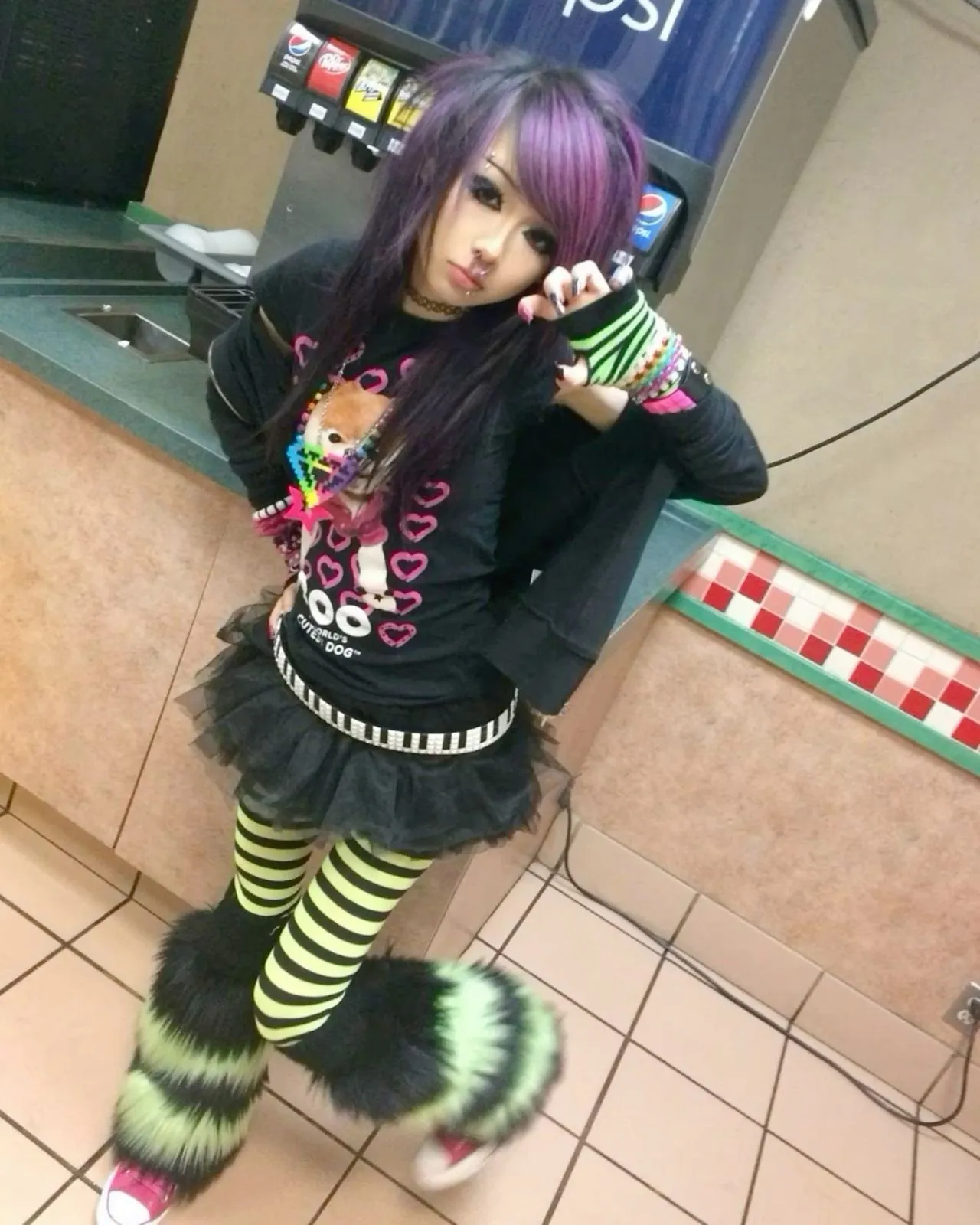 Scene aesthetic — The image perfectly embodies the Scene aesthetic with its heavily teased and dyed hair featuring purple streaks, heavy black eyeliner, a layered outfit including a graphic tee and tutu skirt, striped stockings, fuzzy leg warmers, and colorful accessories like bracelets and a necklace. This aligns precisely with the description's emphasis on neon colors, bold graphics, and the overall mid-2000s MySpace-era look.