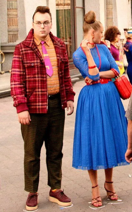 Stilyagi aesthetic — The male figure's pomaded quiff, loud tie, high-collared jacket, and the female figure's bright blue dress and bold accessories directly evoke the Stilyagi aesthetic.