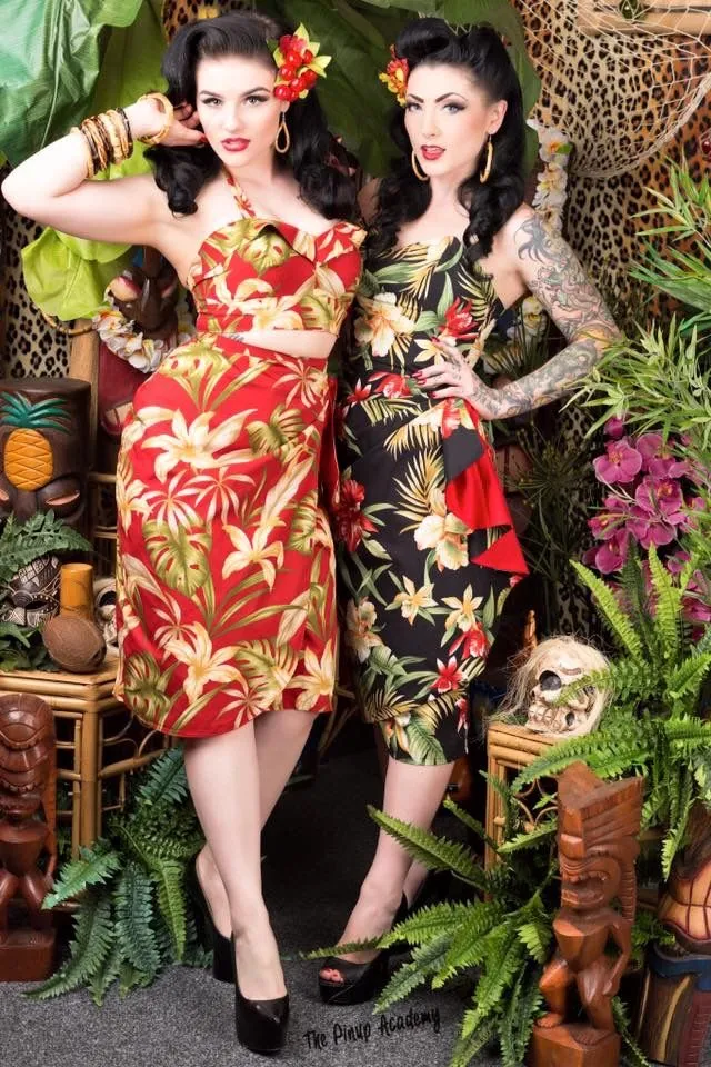 Tiki aesthetic — The two women are dressed in classic Tiki print dresses and styled with hibiscus flowers and bamboo accessories, set against a backdrop of tiki idols and tropical foliage, perfectly capturing the aesthetic.