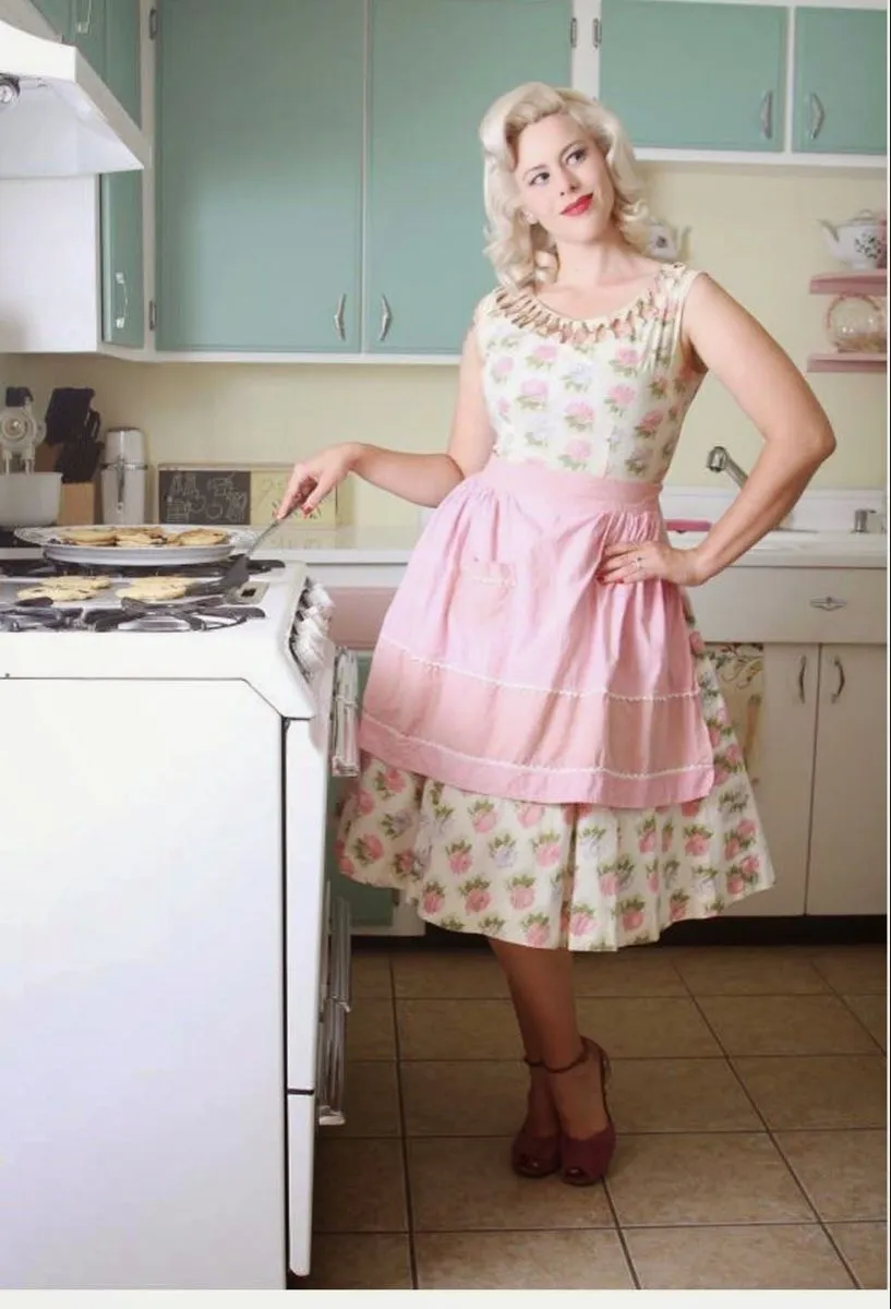 Tradwife aesthetic — The floral dress, embroidered apron, and 1950s housewife pose in a kitchen are direct visual cues for the Tradwife aesthetic.