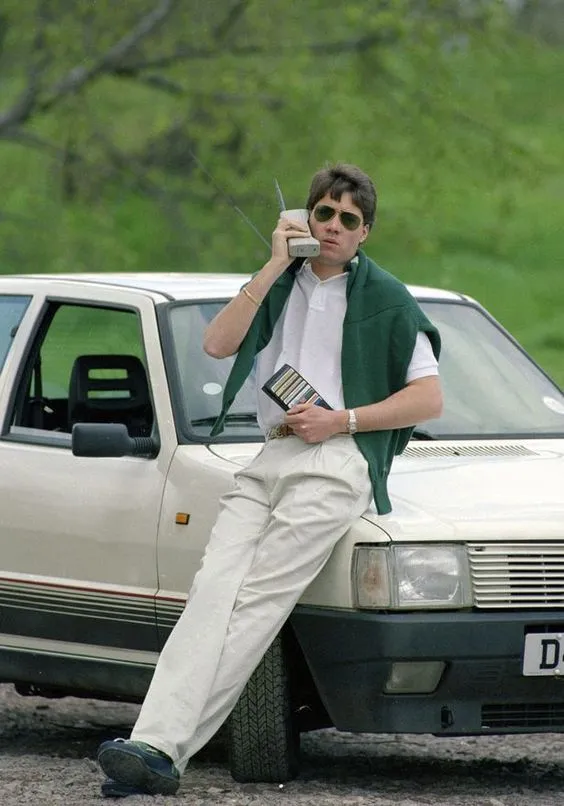 Yuppie aesthetic — The image perfectly captures the 'off-duty' Yuppie look with a Lacoste polo, pleated khakis, a sweater tied around the shoulders, and a car that evokes the era.